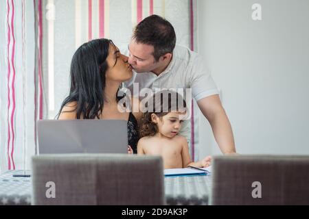 Hispanic mother and Caucasian father kiss happily with their baby girl on their lap painting on the notebook, with a laptop in front of them, sitting Stock Photo