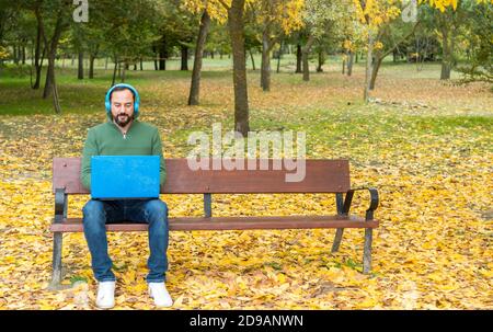 young bearded hipster man sitting on a bench wearing blue wireless headphones while working with his laptop in a park in autumn with fallen tree leave Stock Photo