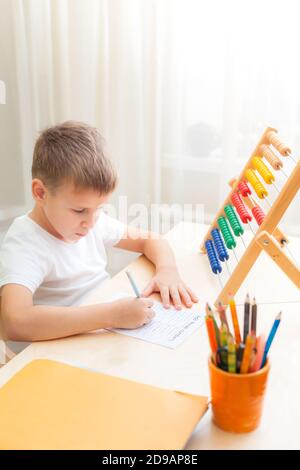 Side view of child solving maths exercises. 7 years old child doing maths lessons sitting at desk in his room. Stock Photo