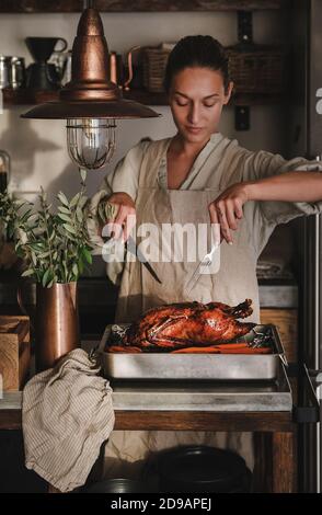 Young woman in apron carving whole oven roasted duck for Thanksgiving holiday or Christmas party celebration in kitchen interior. Traditional Autumn or Winter holiday comfort food concept Stock Photo