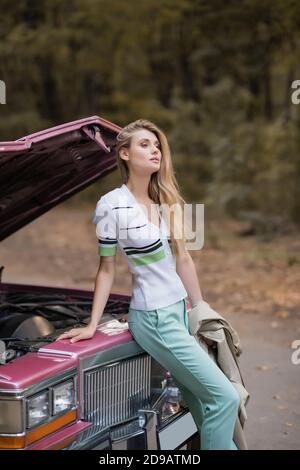 young stylish woman looking away while standing near broken retro car with open hood Stock Photo