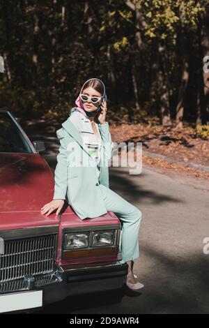 Stylish woman in sunglasses and headscarf sitting on bumper of car near road Stock Photo