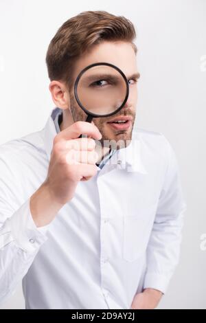 Curious ophthalmologist in white coat looking through magnifying glass isolated on white Stock Photo