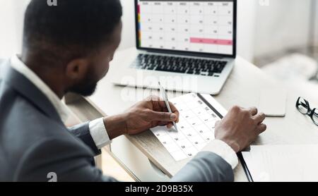 Black Businessman Using Calendar Program On Laptop And Writing Notes To Planner Stock Photo