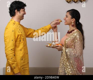 Indian smart couple eating sweet laddu on karva chauth Diwali or anniversary, selective focus. while girl or wife holding puja thali. Stock Photo