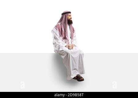 Saudi man in a thobe sitting on a blank panel and looking to the side isolated on white background Stock Photo