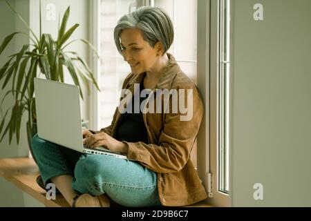 Smiling mature woman looks at web camera taking part in virtual video chat or working with computer or while sitting with her back against window Stock Photo