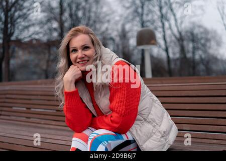 Mature Blonde Woman Smiling And Sitting On Bench In Modern Park After Active Exercising Outdoors. Lifestyle Of Senior Aged People. Sport And Relaxatio Stock Photo