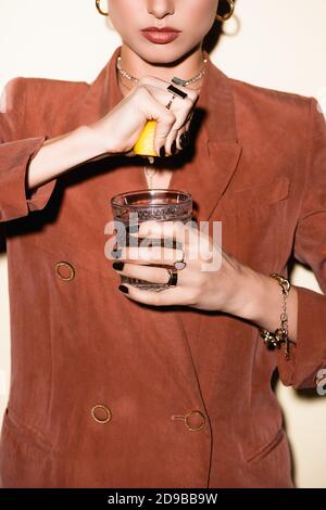 cropped view of young woman in brown blazer squeezing lemon in glass with alcohol cocktail on white Stock Photo
