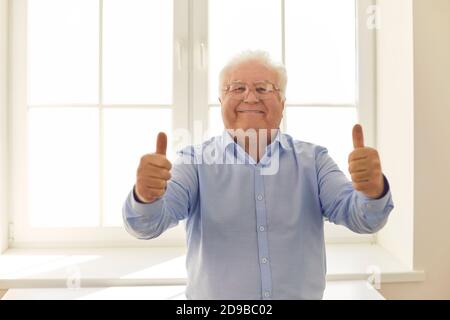 Happy senior man giving thumbs-up and looking at camera standing near the window Stock Photo