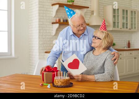 Happy loving senior couple in party hats having quiet birthday celebration at home Stock Photo