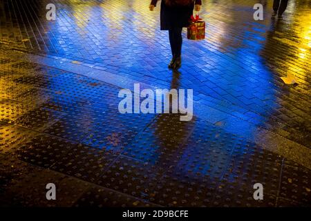 A pedestrian carries a boxed panetone Italian Christmas cake as she crosses the brick paved road illuminated with neon signs after a rain sower had left the roads wet outside the Old Vic Theatre near Waterloo Station in London as the Christmas season is in full-swing. 17 December 2015. Photo: Neil Turner Stock Photo