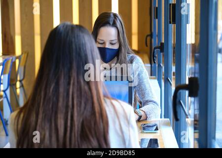 Two female students wearing face masks working on their computers in a restaurant. New normal in restaurants. Coronavirus pandemic.