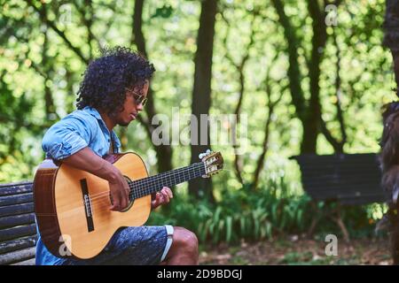 Musician playing guitar in a nice park. He is surrounded by vegetation. Stock Photo