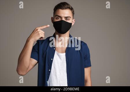 Image of caucasian guy in face mask pointing finger at his face isolated over grey background Stock Photo