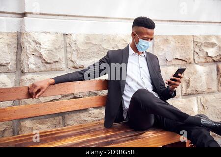 Businessman Wearing Mask Sitting Outdoors On Bench With Phone Having Lunch Break During Pandemic Stock Photo