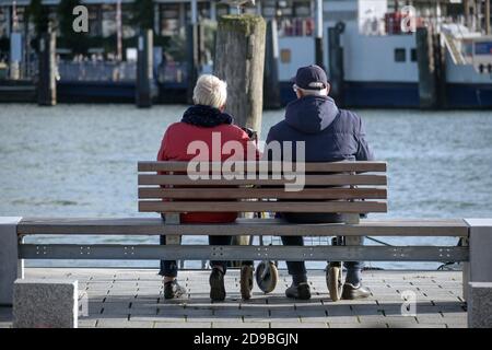 Older couple from behind sitting on a bench directly on the waterfront on the quay and enjoying the port life in the sun, selected focus Stock Photo
