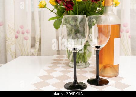 Two glasses and bottle of wine on table with roses flowers in vase as romantic date or celebrating anniversary concept Stock Photo