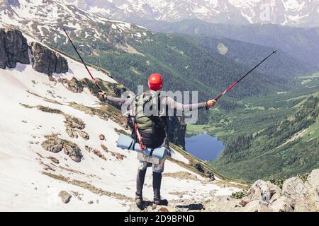 Happy tourist with large backpack and sticks climbed to top of mountain and rejoices at achieved goal by raising hands up against background of high s Stock Photo