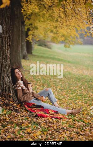 Beautiful woman in sweater, jeans and boots sitting under the maple tree with cup of coffee or tea in autumn park. Stock Photo