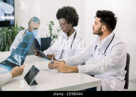 Cropped image of team of multiethnic doctors, looking doctor speech on online video conference at light room. Focus on handsome bearded Indian male Stock Photo