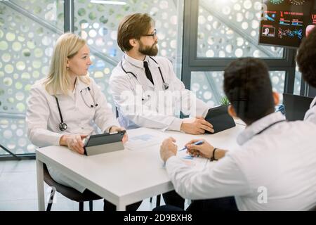 Cropped side angle view of multi-ethnic group of young doctors sitting around table during medical staff meeting or conference in clinic, and looking Stock Photo