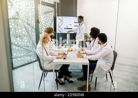 Handsome African medical doctor using big digital screen and charts, having a lecture report for his multiethnic colleagues, sharing the results of Stock Photo