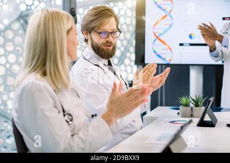 Close up of two Caucasian male and female doctors, sitting at the table in meeting room, and clapping for African colleague after successful Stock Photo