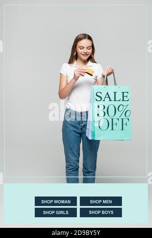 joyful woman holding credit card and looking at blue shopping bags with sale 30 percent off lettering on grey Stock Photo