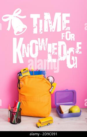 yellow backpack with school supplies, lunch box, toy school bus and pen holder with felt pens near time for knowledge lettering on pink Stock Photo
