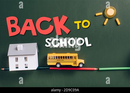 top view of toy school bus on road made of color pencils, house model and sun on green chalkboard with back to school lettering Stock Photo