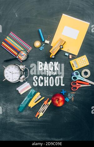 top view of frame with vintage alarm clock, green apple and school supplies on black chalkboard with back to school lettering Stock Photo