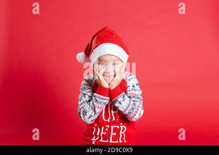 close up portrait of smiling and surprised girl in Santa Claus hat holding hands to cheeks, isolated on red background Stock Photo