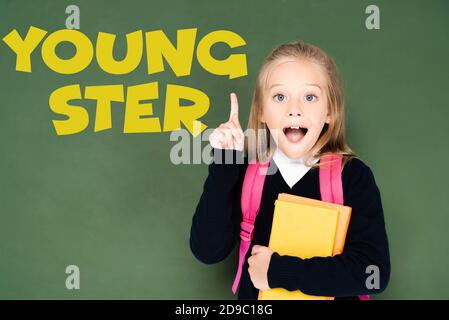 excited schoolgirl holding book and showing idea gesture while standing near green chalkboard with youngster illustration