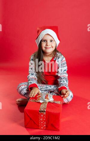 a girl in pajamas and a Santa Claus hat is sitting barefoot on the floor next to a Giftbox on a red background, isolated Stock Photo