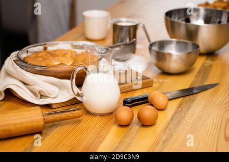 Tasty apple pie on a oaken wood table with a ingredients. Woman in a gray pajama cooking a delicious apple pie on a kitchen in a loft style. Milk Stock Photo