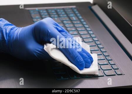 A mans hand in a blue glove cleans a laptop keyboard with an antibacterial agent to protect against the COVID-19 epidemic. Cleaning the surface from b Stock Photo
