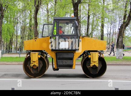 The asphalt rink is yellow with iron wheels on the road in the city against the background of green trees. Repair work on the street on laying asphalt. Stock Photo
