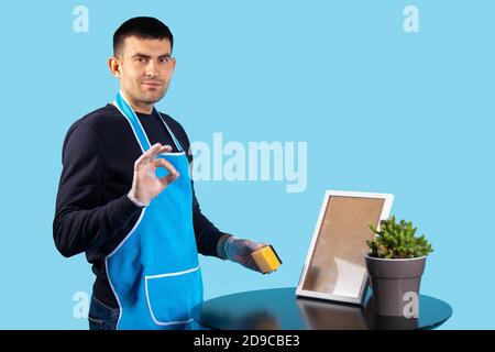 A handsome man in an apron and gloves, wipes off his ardor and looks contentedly at the camera. Photo on a blue, ok sign. Gender stereotypes concept Stock Photo