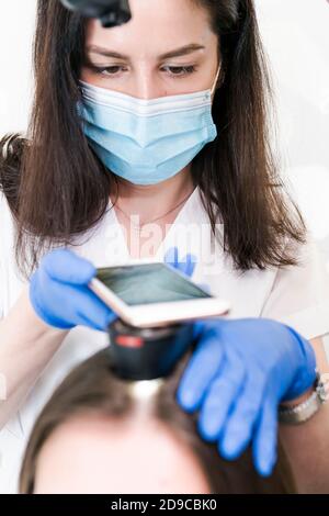 The doctor dermatologist examining client hair with a special apparatus for diagnostic hair. Trichoscopy procedure in the trichology clinic. Concept o Stock Photo