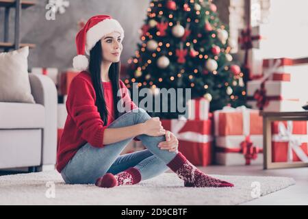 Photo of young charming girl sit carpet embrace knee wear santa headwear red pullover jeans socks in decorated living room indoors Stock Photo
