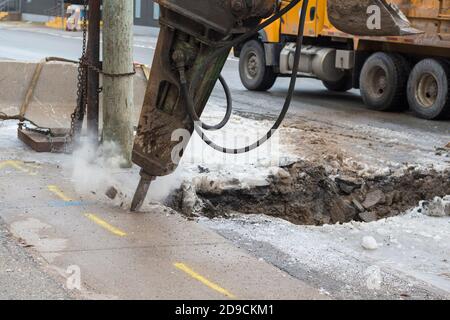 A large pneumatic jackhammer breaking a frozen road and sidewalk. Bits of concrete fly from the point. Stock Photo