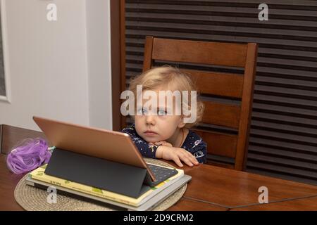 Little tired girl child uses a tablet sitting at the table. Childhood with modern technology. Modern childhood. Playing and learning with the help of Stock Photo