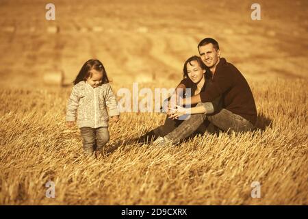 Happy young father and mother with two year old baby girl sitting on the ground in harvested field Stock Photo
