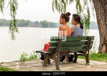 Two women relaxing sitting peacefully on park bench. Asian mom and daughter tourist at Beijing summer palace enjoying quiet summer day traveling in Stock Photo