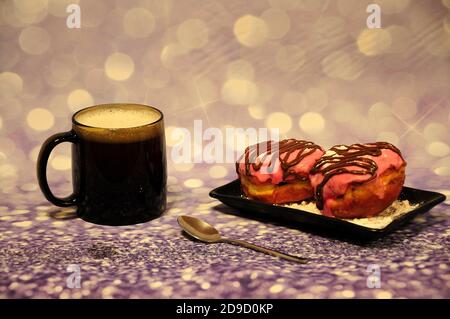 Two donuts in pink glaze and decorated with chocolate and powdered sugar on a plate next to a mug of cappuccino on a gray shiny background. Close-up. Stock Photo