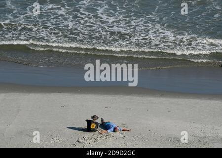 Two men on beach one overweight laying stretched out other sitting next to him Stock Photo