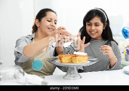 Hispanic mom and daughter decorating a cake with whipped cream - Latin mom teaching her daughter to decorate a cake - Mother and daughter having fun c Stock Photo