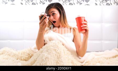 woman rises in bed with a cup of coffee and an alarm clock in her hands, in horror realizes that she overslept. Concept of lack of time, being late Stock Photo