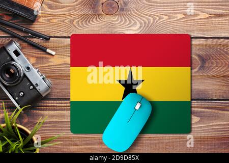 Ghana flag on wooden background with blue wireless mouse on a mouse pad, top view. Digital media concept. Stock Photo